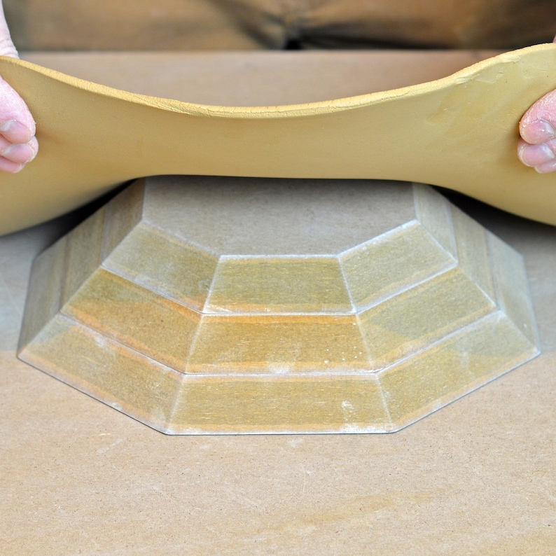 May include: A light brown ceramic bowl with a multi-tiered, octagonal base. The bowl is being held by hands, with a thin sheet of clay covering the top.