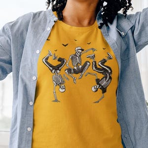 May include: Mustard yellow t-shirt with a graphic of three dancing skeletons and bats. The t-shirt is worn with a light blue button-down shirt and blue jeans. The person is wearing headphones and holding a yellow phone.