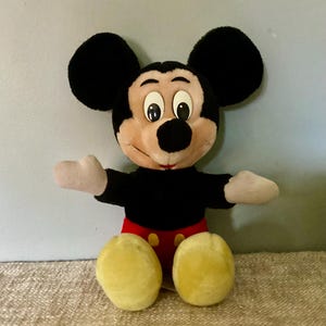 May include: A vintage plush Mickey Mouse toy with black fur, large ears, and a red and yellow outfit. The toy has a friendly expression with big eyes and a wide smile.