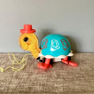 May include: A vintage pull-along toy turtle with a yellow head, a turquoise shell with red and white accents, and red legs. The turtle wears a red hat and has a yellow pull string.