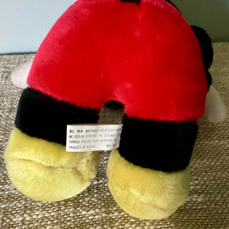 May include: A red, black, and yellow plush toy shaped like a cartoon character. The toy has a tag that reads "ALL NEW MATERIALS-POLYESTER FIBER", "ME 1525-PA 1378 OHIO PA 1378 MASS", "CANASA TRADING CORP, BURBANK, CA", "PRODUCT OF KOREA", and "MR-."