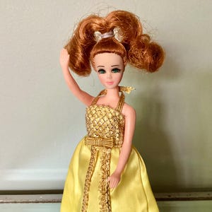 May include: A vintage doll with red hair styled in two high ponytails, adorned with a silver bow. The doll wears a yellow satin gown with gold accents and a gold bodice. The doll has green eyes and is holding up one arm.