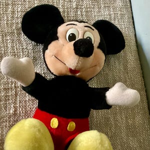 May include: A plush Mickey Mouse toy with black fur, large ears, and a red and yellow outfit. The toy has a friendly expression with big eyes and a wide smile.