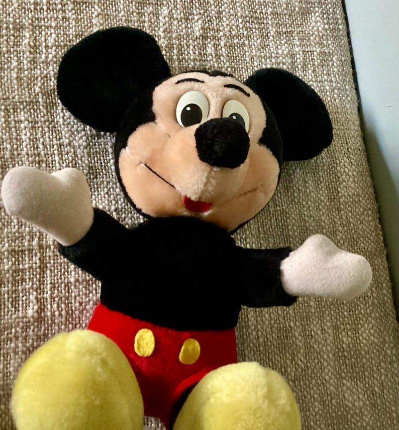 May include: A plush Mickey Mouse toy with black fur, large ears, and a red and yellow outfit. The toy has a friendly expression with big eyes and a wide smile.