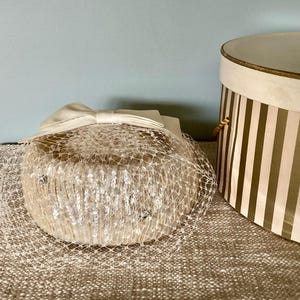 May include: A white hat with a veil and a satin bow. The hat is sitting on a brown surface.