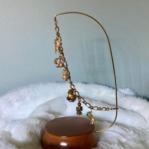 May include: A gold-toned charm bracelet with various charms, including a crown, owl, and boot, hangs from a curved gold stand. The stand is mounted on a round, wooden base. The background is a soft, white, fluffy material.