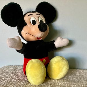 May include: A plush Mickey Mouse toy with black ears, a black body, red shorts, and yellow shoes. The toy is sitting on a brown and white textured surface.