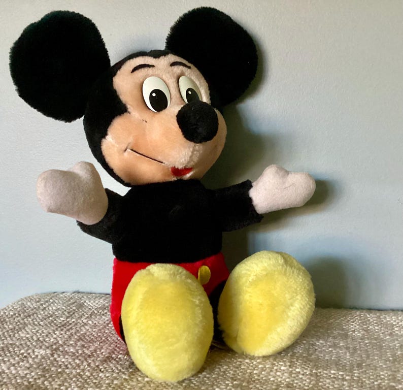 May include: A plush Mickey Mouse toy with black ears, a black body, red shorts, and yellow shoes. The toy is sitting on a brown and white textured surface.