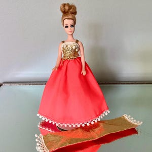 May include: A vintage doll in a coral-colored gown with a gold bodice and a matching gold and white decorative piece. The doll has blonde hair styled in a bun. The dress has a full skirt and is trimmed with white pom-poms.