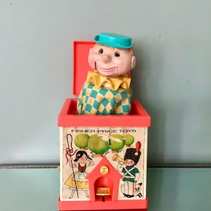 May include: A vintage Fisher-Price jack-in-the-box toy featuring a plastic clown with a teal hat and a yellow ruffled collar emerging from a red square box. The box sides display illustrations of a girl with a staff and a soldier with a trumpet.