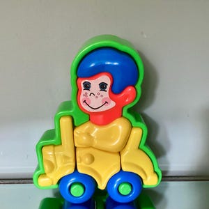 May include: A colorful, vintage toy featuring a smiling monkey face with blue hair and red ears. The monkey is set on a yellow body with green and blue wheels. The toy is made of plastic and is designed for children.