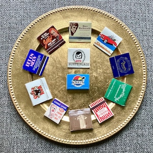 May include: A collection of vintage matchbooks featuring various businesses and locations. The matchbooks are arranged on a gold-colored plate with a beaded edge. Some of the matchbooks feature images of people, while others feature text and logos. The matchbooks are in various colors, including red, blue, green, and white.