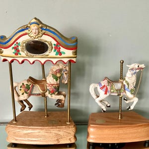May include: Two carousel horse figurines. One is under a decorative canopy with a mirror, the other is a single horse. Both are white with floral designs, brown saddles, and gold accents. They sit on wooden bases.