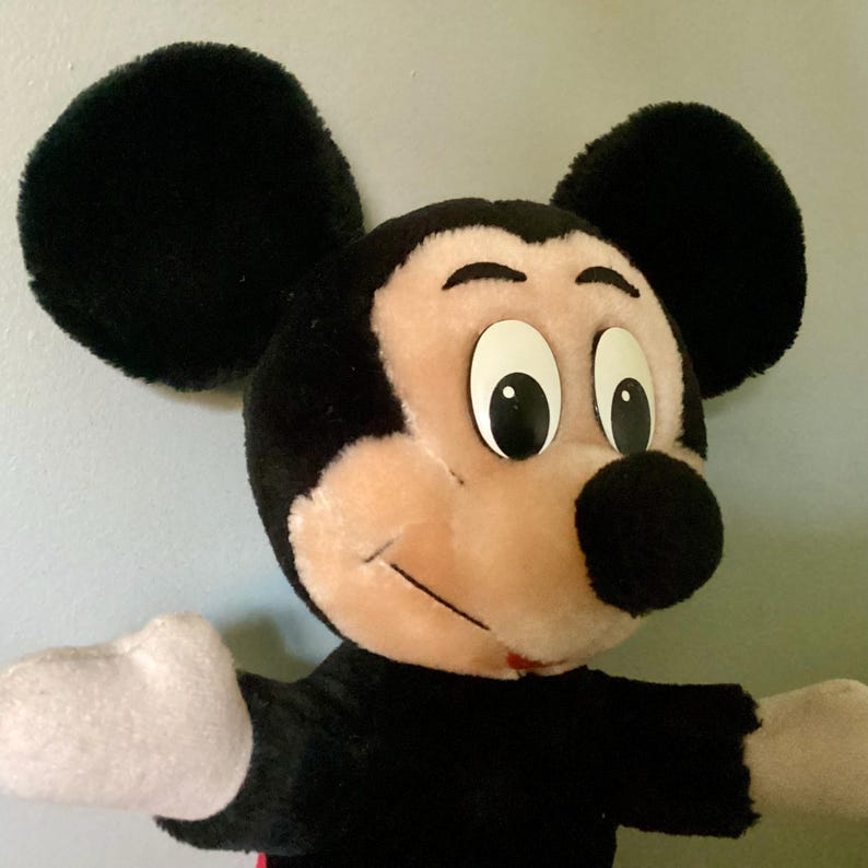 May include: A plush Mickey Mouse toy with black fur, large black ears, and a pink face. The toy has large black eyes with white pupils and a black nose.