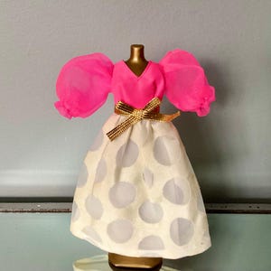 May include: A miniature dress on a gold mannequin. The dress features a bright pink top with puffy sleeves and a V-neck. The skirt is cream-colored with large, light gray polka dots. A gold ribbon belt cinches the waist, adding a touch of elegance.