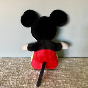 May include: A plush Mickey Mouse toy with black fur, red shorts, and a black tail. The toy is sitting on a beige surface.