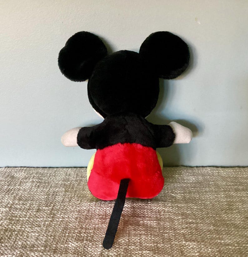 May include: A plush Mickey Mouse toy with black fur, red shorts, and a black tail. The toy is sitting on a beige surface.