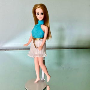 May include: A doll with long blonde hair, blue eyes, and painted facial features. The doll wears a sleeveless turquoise top with a high neck and a white pleated skirt featuring a thin, metallic gold belt. The doll stands on a circular base.