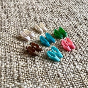 May include: A collection of miniature doll shoes in various colors, including white, brown, teal, green, and pink. The shoes are arranged on a textured, neutral-toned surface. The shoes appear to be made of plastic.
