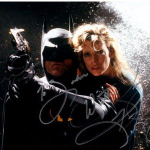 Può includere: Un costume di Batman nero e grigio con un mantello nero e una donna con una camicia blu. La donna tiene il braccio di Batman e entrambi guardano la telecamera.