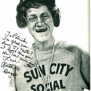 May include: Black and white photo of a woman wearing a grey sweatshirt with the text "SUN CITY SOCIAL DIRECTOR" on it. She is smiling and has a whistle around her neck. The woman has signed the photo with the message "Jo Patrick, I'm glad you have My Taste!! Love this pie! Ha! Ha! Love - Ruthie Buzze '74"