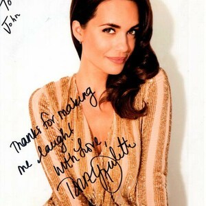 Può includere: Una donna con lunghi capelli castani indossa un vestito a righe dorate e bianche. Sorride e guarda la telecamera. Il testo "To John Thanks for making me laugh! With love,  " è scritto in inchiostro nero su uno sfondo bianco.