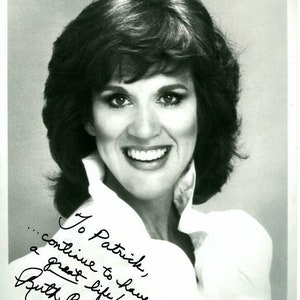 May include: Black and white photo of a woman with short brown hair smiling. She is wearing a white collared shirt. The photo is signed "To Patrick, ...continue to have a great life! Olsay?  Ruth Buzzi"