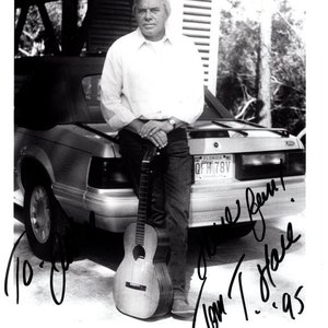Op de afbeelding: Zwart-witfoto van Tom T. Hall, een countrymuzikant, die naast een auto staat met een gitaar in zijn hand. Hij draagt een wit overhemd en een spijkerbroek. De foto is gesigneerd door Tom T. Hall en bevat de tekst "World Class Talent".