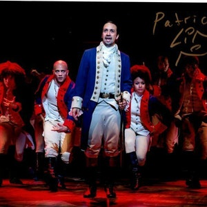 May include: A group of actors dressed in red, white, and blue costumes, performing on a stage. The actor in the center is holding a microphone and singing. The signature "Patrick Page" is written in gold ink in the top right corner of the image.