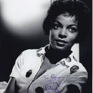 Könnte beinhalten: Schwarzweißfoto einer Frau mit kurzen, dunklen Haaren. Sie trägt ein weißes Hemd mit einem Tupfenmuster. Das Foto ist signiert mit "To Ann-Joy,  Ruby Dee".