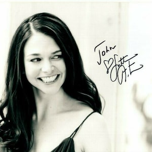 May include: Black and white photograph of a woman with long dark hair. She is smiling and looking to the right. The photo is signed "John Slattery" with a heart symbol.