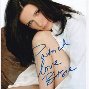 Puede incluir: Una mujer con el pelo largo y oscuro lleva una camisa blanca y tiene el brazo apoyado en la rodilla. La imagen está firmada "Patrick Love Bitsie".
