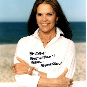 以下が含まれることがあります： 白いシャツに赤いネックレスとブレスレットを着けた女性。彼女は微笑んでカメラを見ている。背景は青い水と白い砂浜のビーチ。女性はシャツに「For John, Best wishes & Peace -  A. MacGraw」というメッセージを書いてサインしている。