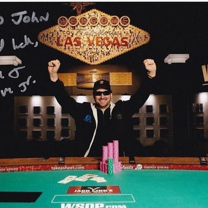 Pode incluir: Fotografia de um homem celebrando uma vitória no pôquer em Las Vegas. Ele está usando óculos de sol e uma jaqueta preta, com os braços levantados. O fundo apresenta a placa "Welcome to Las Vegas" e fichas de pôquer.