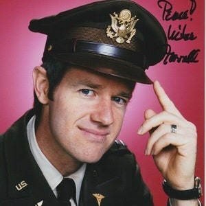 May include: A man wearing a military uniform with a black cap and a gold eagle insignia. He is smiling and holding his cap with his right hand. The inscription "To Team - Peace!  Love,  Maxwell" is written in black ink on a pink background.
