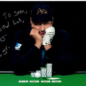 Pode incluir: Fotografia autografada de um jogador de poker com um boné preto com a inscrição "Aria" em dourado. O jogador apoia a cabeça na mão, com uma luva branca. Fichas e cartas de poker sobre uma mesa de feltro verde. Texto manuscrito.