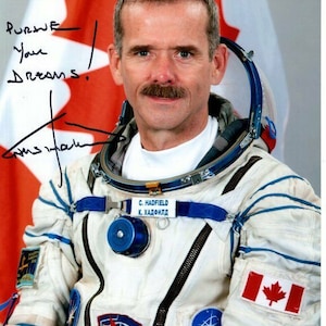 Puede incluir: Un astronauta canadiense con un traje espacial blanco con detalles azules y un parche de la bandera canadiense en la manga. El astronauta lleva un casco y tiene bigote. El astronauta está de pie frente a una bandera canadiense. El texto "Pursue your dreams." está escrito con tinta negra en la imagen.