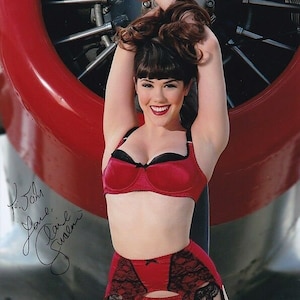 May include: A woman wearing a red satin bra and matching garter belt with black lace trim. She is posing in front of a red airplane propeller. The image is signed "To John, Love, Claire Sinclair".