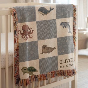 May include: A gray and cream baby blanket with a sea life theme, featuring a whale, octopus, dolphin, seal, and turtle. The blanket has a checkered pattern and a fringed edge. The name "OLIVER" and the date "14 AUG 2026" are printed on the blanket.