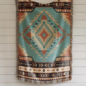 May include: A woven throw blanket featuring a large central diamond pattern in shades of teal, rust orange, cream, and dark brown. The design includes geometric shapes and arrow motifs with a fringed edge along the top and bottom borders.