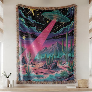May include: A woven tapestry featuring a desert landscape with cacti, mountains, and a starry sky. A UFO emits a pink beam of light. The tapestry has a fringed edge and is in shades of pink, blue, and green.