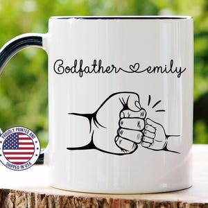 May include: White 11 oz ceramic mug with a black handle and rim. The mug features the text "Godfather emily" in a script font, along with a graphic of two fists bumping. A small American flag sticker is visible.