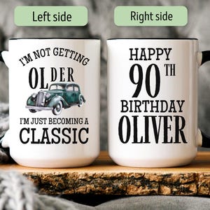 May include: A white ceramic mug with a black handle and rim, featuring a 15 oz capacity. The left side displays a green vintage car graphic with the text I'M NOT GETTING OLDER I'M JUST BECOMING A CLASSIC. The right side reads HAPPY 90TH BIRTHDAY OLIVER.