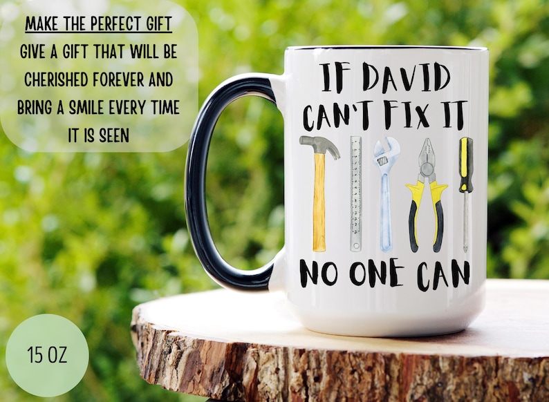 Op de afbeelding: Een witte keramische mok met een zwart handvat. De mok heeft een zwarte afbeelding met de tekst "Als David het niet kan repareren, kan niemand het" met een tekening van gereedschap, waaronder een hamer, een liniaal, een moersleutel, een tang en een schroevendraaier.
