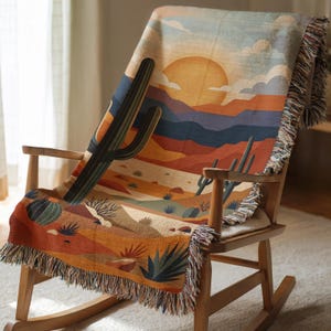 May include: A woven throw blanket featuring a desert landscape scene with a large orange sun, layered mountains, and various cacti. The blanket has a fringed edge and is draped over a wooden rocking chair, displaying warm tones of orange, blue, and tan.