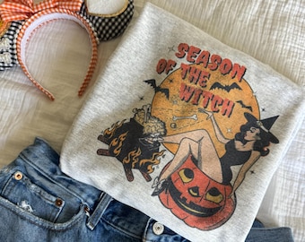 May include: A light grey sweatshirt with the text "SEASON OF THE WITCH" and a Halloween graphic, alongside denim shorts and a Halloween headband with orange and black details.