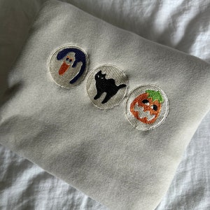May include: A light gray sweatshirt with three embroidered patches. The patches feature a blue ghost, a black cat, and an orange jack-o'-lantern. The sweatshirt is folded on a white surface.