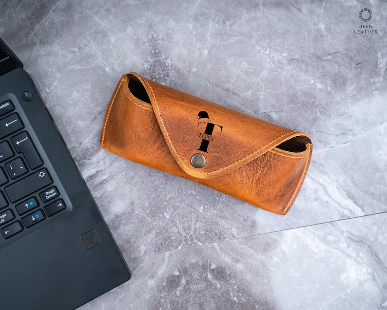 May include: A tan leather glasses case with a press stud closure. The case features the name "TONY" and a stylized "T" printed on the front. The case is on a grey marble surface next to a black laptop.