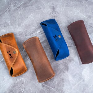 May include: Four leather eyeglass cases in tan, brown, blue, and burgundy. The tan case has the initials "J A T" and a heart symbol. The blue case has the initials "S K". Each case has a button closure.