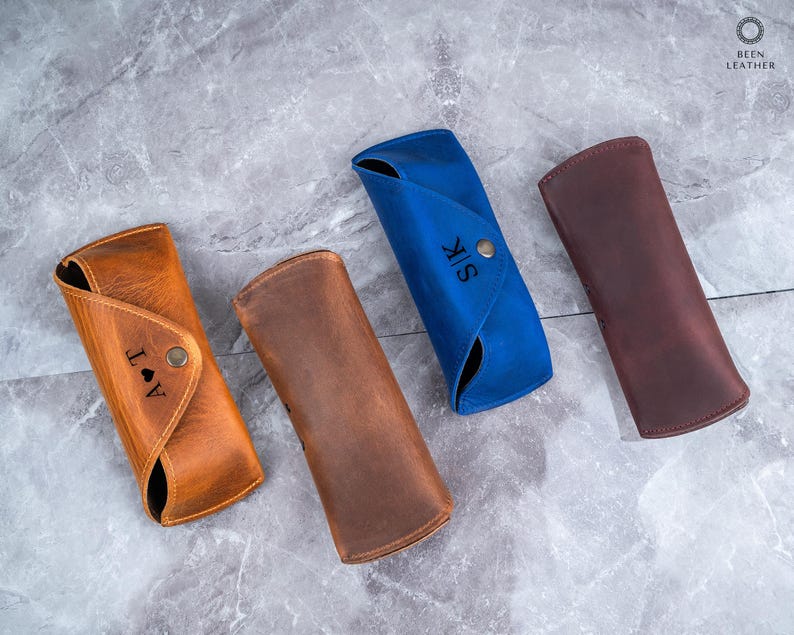 May include: Four leather eyeglass cases in tan, brown, blue, and burgundy. The tan case has the initials "J A T" and a heart symbol. The blue case has the initials "S K". Each case has a button closure.